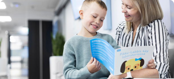 Lapsi istuu äitinsä sylissä laboratoriossa ja lukee Fimlabin Pikku-Labbis-kirjasta.